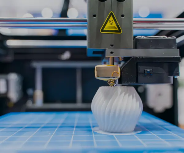 3D printer using FDM technology to create a white plastic vase layer by layer on a blue build platform.
