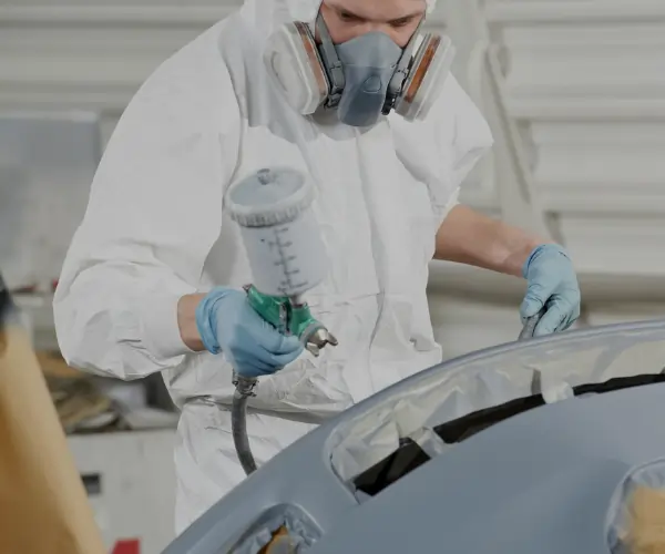 Worker in safety gear spray painting a car part with precision using a professional paint sprayer.