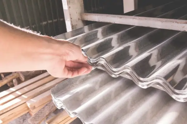 A hand touching a stack of corrugated metal sheets with a shiny, protective zinc plating.