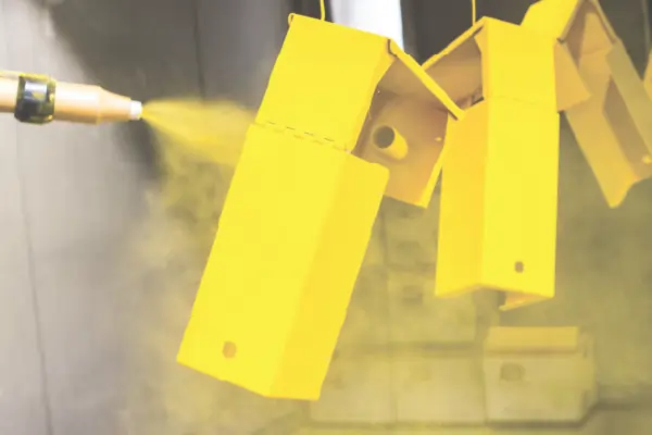A worker applying a yellow powder coat to several metal parts hanging in a spray booth.